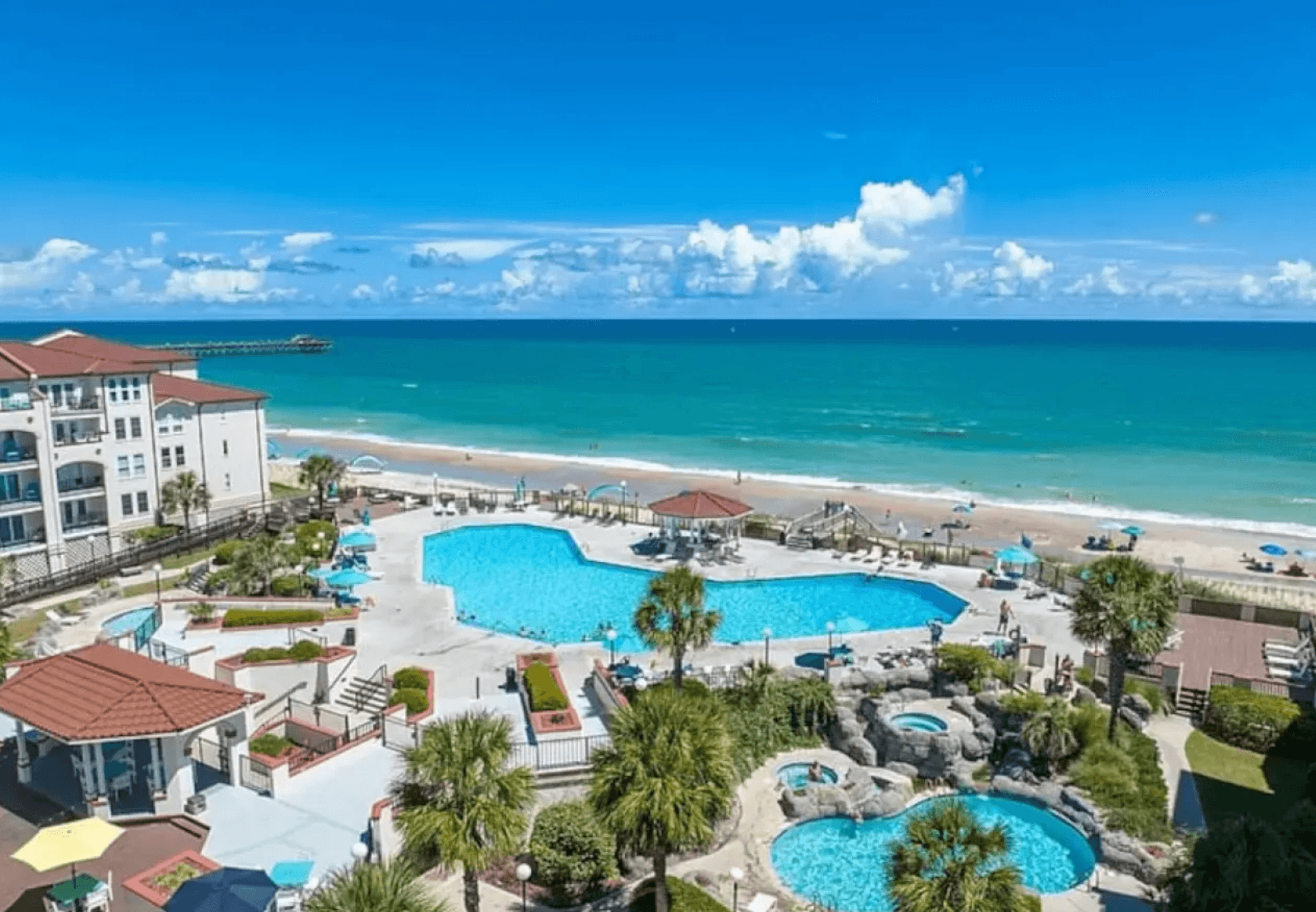 The Villa Capriani pool complex with palm trees, hot tubs, and the Atlantic beach and pier beyond, North Topsail Beach