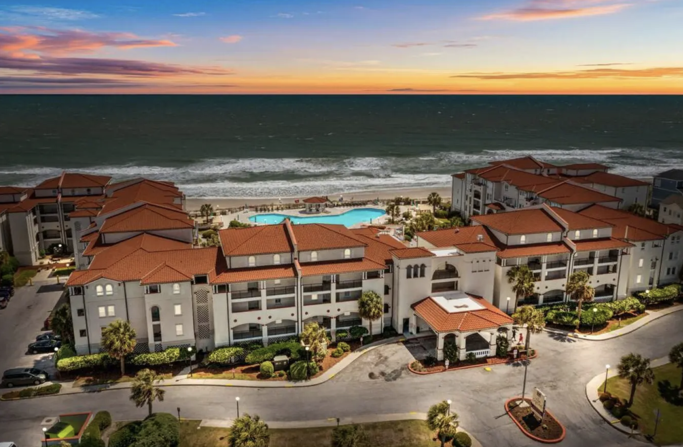 Aerial view of Villa Capriani's salmon-pink Mediterranean buildings and terracotta-tile roofs at sunset with the Atlantic behind, North Topsail Beach