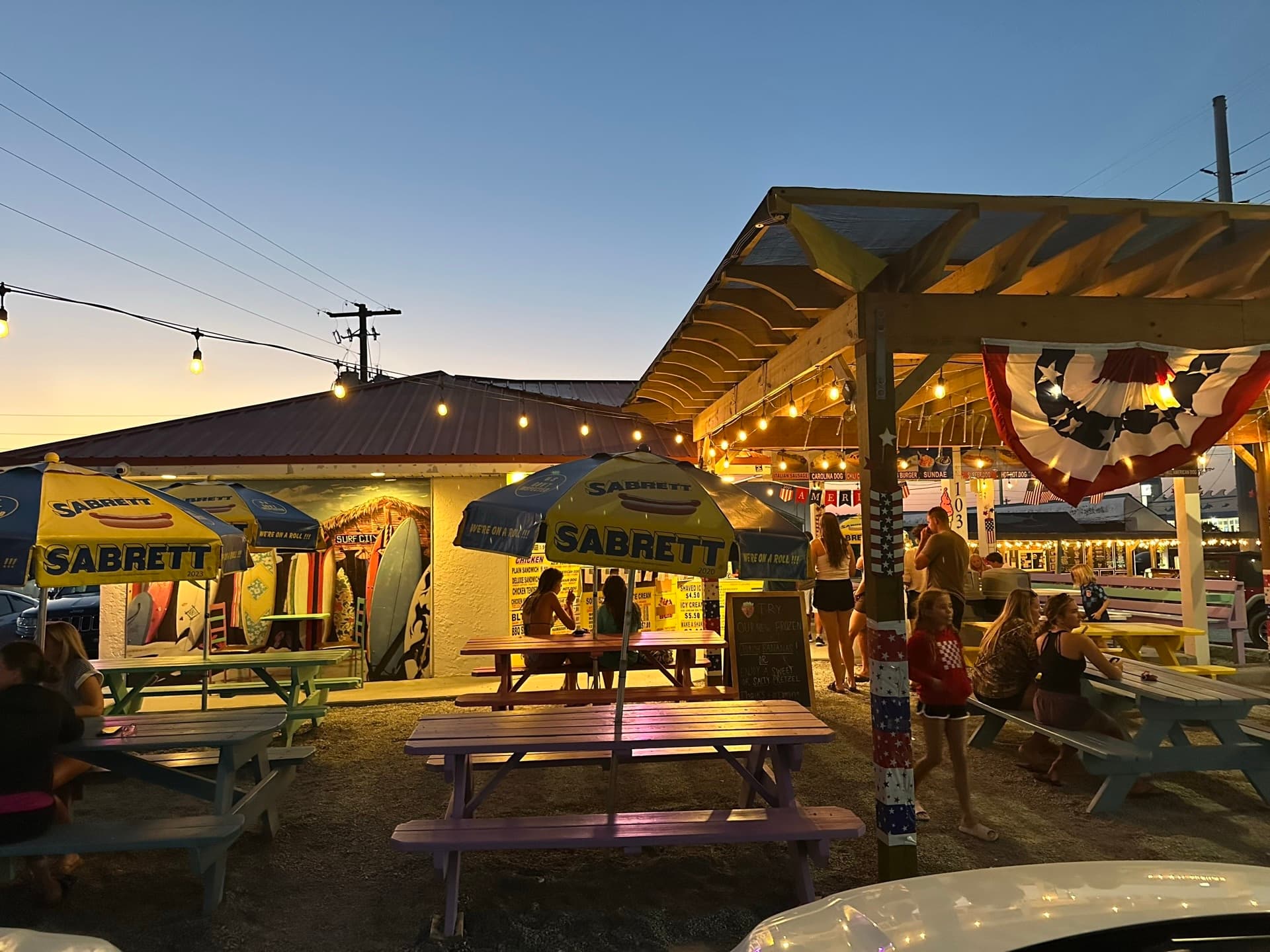 Hot Diggity Dogz open-air ice cream and hot dog stand in Surf City at dusk, string lights glowing over colorful picnic tables